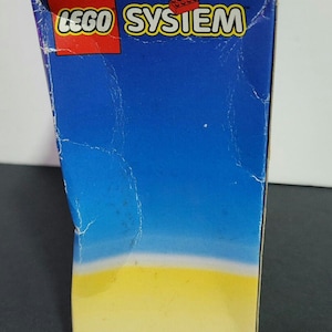Puede incluir: Una caja azul y amarilla con las palabras "LEGO SYSTEM" impresas en rojo en la parte delantera.