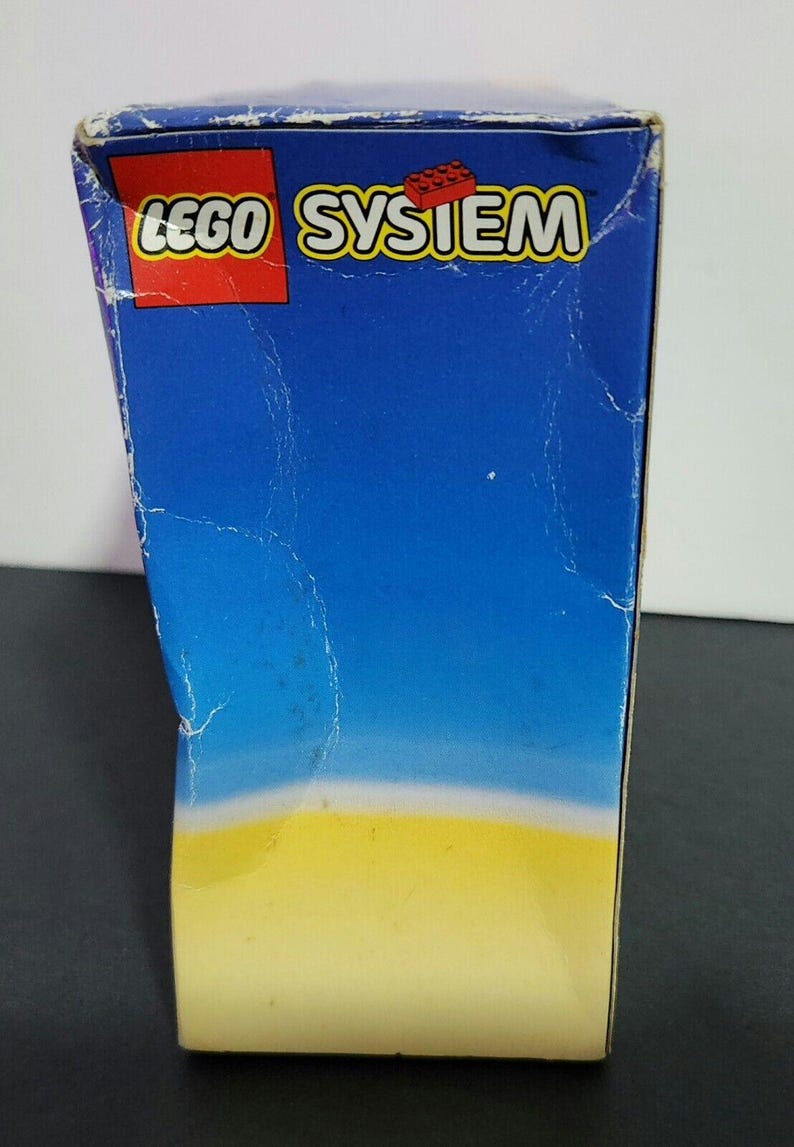 Puede incluir: Una caja azul y amarilla con las palabras "LEGO SYSTEM" impresas en rojo en la parte delantera.
