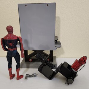 Może przedstawiać: Figurka Spider-Mana w czerwono-czarnym kostiumie stoi obok szarej, prostokątnej konstrukcji. Figurka ma na plecach emblemat pająka. Obecne jest r&oacute;wnież czarne urządzenie mechaniczne i małe srebrne elementy.