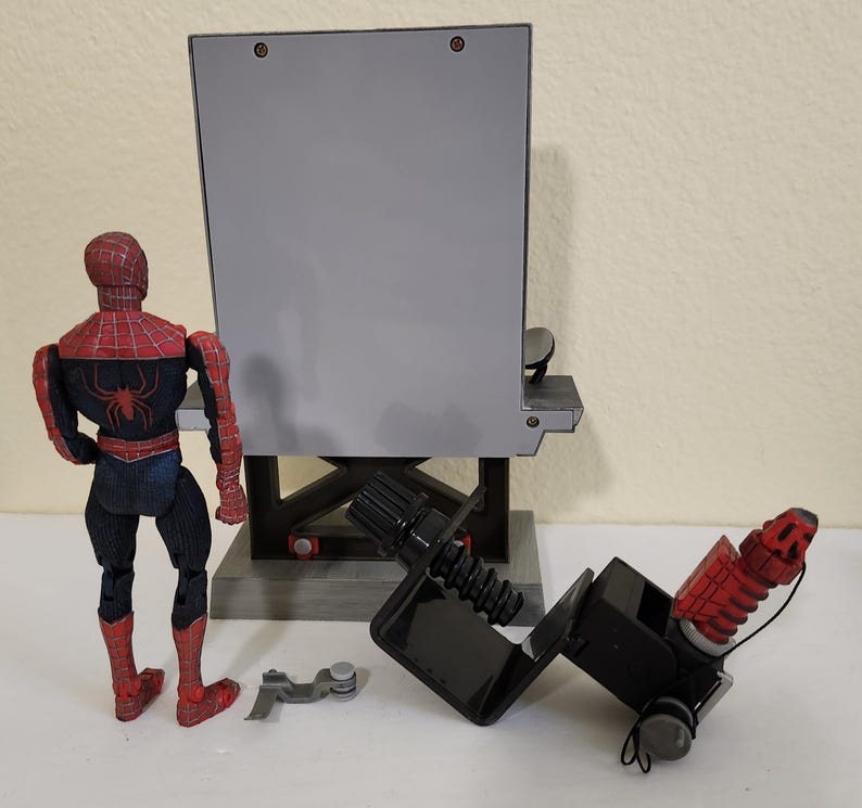 Może przedstawiać: Figurka Spider-Mana w czerwono-czarnym kostiumie stoi obok szarej, prostokątnej konstrukcji. Figurka ma na plecach emblemat pająka. Obecne jest r&oacute;wnież czarne urządzenie mechaniczne i małe srebrne elementy.