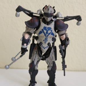 Pode incluir: Figura de ação detalhada de um guerreiro em armadura de batalha. A figura é principalmente cinza, com detalhes em preto e vermelho. Possui capacete, ombreiras e várias armas. O peito apresenta um emblema azul. A figura está em pé.