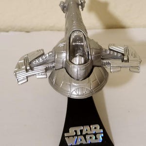 May include: A silver-colored Star Wars spaceship model. The model is mounted on a black base with the Star Wars logo in silver. The spaceship has detailed wings and a cockpit.