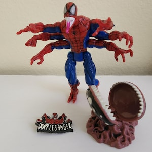 May include: A Spider-Man action figure with six arms, red and blue suit, and a long pink tongue. Includes a small pin with the word "Doppelganger" and a brown, open-mouthed creature.