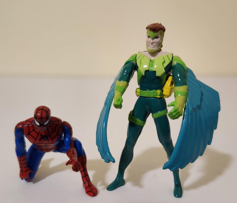 Pu&ograve; includere: Due action figure: Spider-Man in posizione accovacciata e un supereroe con ali verdi e blu. La figura del supereroe indossa un costume verde e blu con cintura e accessori gialli. Le figure sono su una superficie bianca.