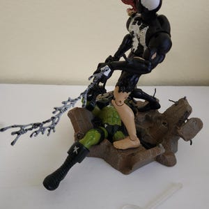 May include: A black and white Venom action figure with a red tongue attacks a green and black action figure. The figures are on a brown base. A clear plastic stand is in the foreground. The figures are from a comic book or movie.