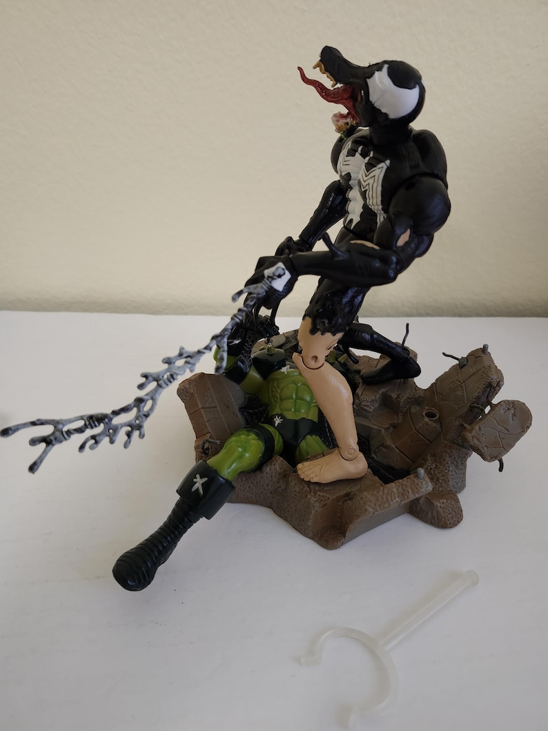 May include: A black and white Venom action figure with a red tongue attacks a green and black action figure. The figures are on a brown base. A clear plastic stand is in the foreground. The figures are from a comic book or movie.