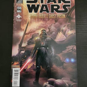 May include: A Star Wars comic book featuring Darth Vader. The cover art shows Darth Vader with a red lightsaber, set against a backdrop of a futuristic structure. The title "Star Wars: Darth Vader and the Ghost Prison" is prominently displayed.