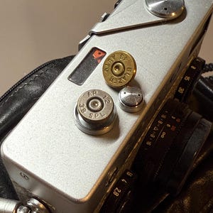 May include: Silver camera with a black lens and leather case. The camera has a circular top and several metal accents. Two bullet casings are on top, one marked "45 AUTO BLAZER" and the other "38 SPL AP".