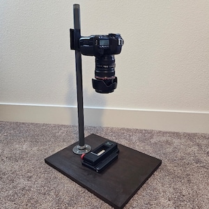 May include: A black camera stand featuring a dark wooden base, a vertical metal pole, and a black camera mount. A black digital camera with a lens and a lens hood is attached to the mount, positioned to face downward toward the base of the stand.