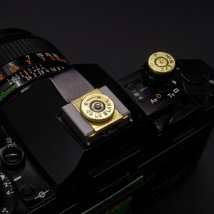 May include: Black camera with brass bullet casing flash shoe cover and hot shoe cap. The camera has a lens with numbers and markings. The flash shoe cover has the text "45 LC BLANK". The hot shoe cap has the text "NIM 243".