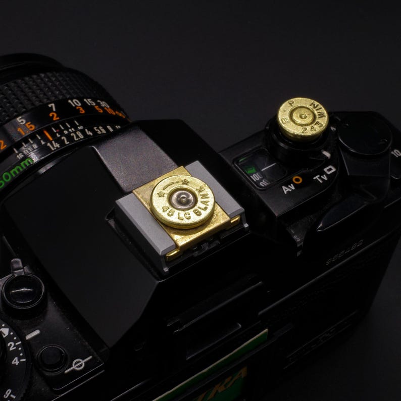May include: Black camera with brass bullet casing flash shoe cover and hot shoe cap. The camera has a lens with numbers and markings. The flash shoe cover has the text "45 LC BLANK". The hot shoe cap has the text "NIM 243".