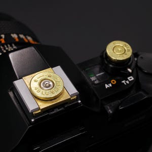 May include: Close-up of a black camera with brass-colored bullet casings used as decorative elements. One casing is on the flash mount, and another is on a dial. The camera has various control settings visible.