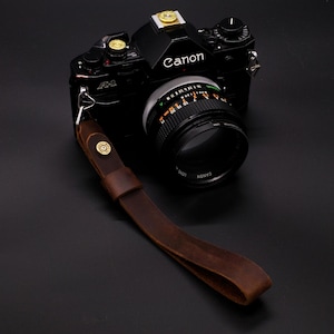 May include: A black Canon A-1 film camera with a brown leather camera strap. The camera has a lens with a Canon logo and a silver ring with markings for aperture and shutter speed.
