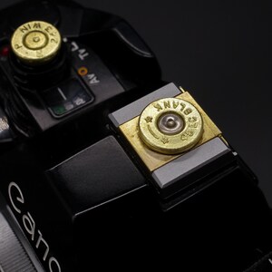 May include: Close-up of a black Canon camera with custom brass bullet casing accents. The casings are engraved with text and set into the camera's controls. The camera has a sleek, modern design.