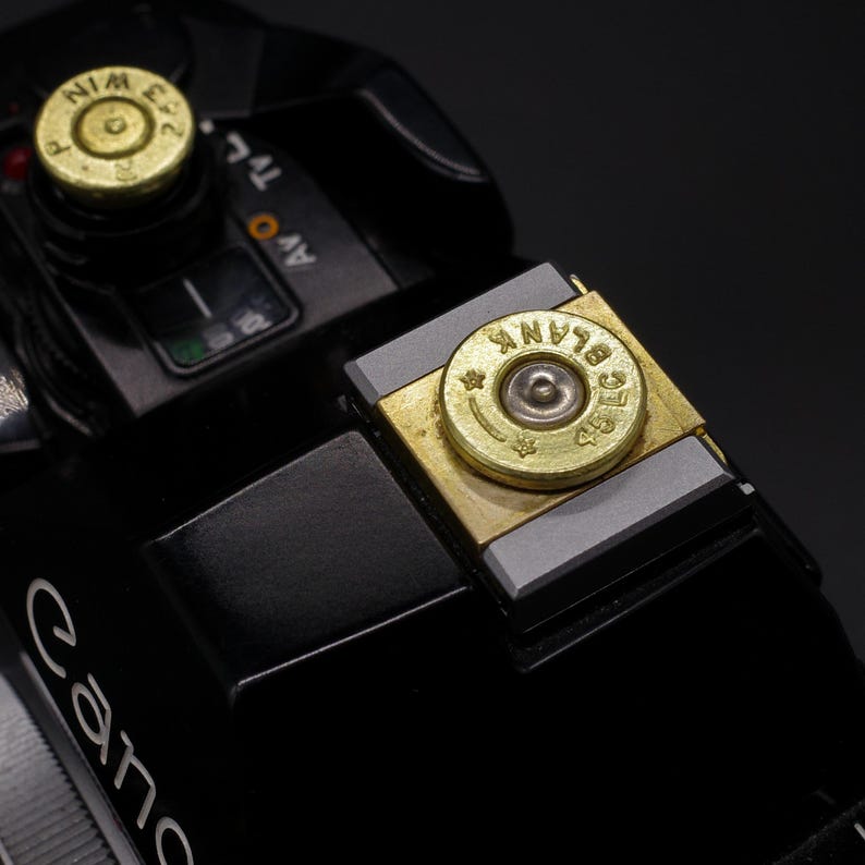 May include: Close-up of a black Canon camera with custom brass bullet casing accents. The casings are engraved with text and set into the camera's controls. The camera has a sleek, modern design.