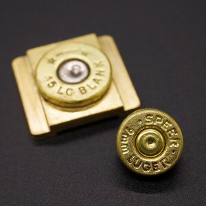 May include: Two brass bullet casings, one with "45 LC BLANK" and the other with "9mm LUGER SPEED" text. One casing is set in a brass holder. The casings are on a dark surface.