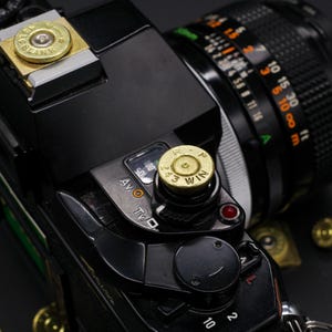 May include: Close-up of a black camera with a lens and brass bullet casings attached. The casings are engraved with text. The camera has various dials and settings visible, including "AV O" and "Tv O".