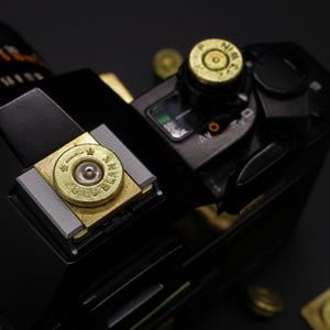 May include: Close-up of a black camera with brass bullet casings. The casings are used as decorative elements on the camera's top and a dial. The camera has a sleek, modern design with various buttons and dials.