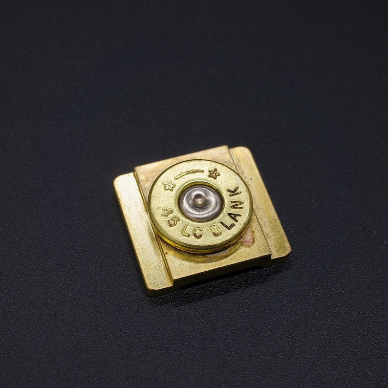 May include: A square, brass-colored money clip with a .45 LC BLANK bullet casing in the center. The bullet casing is gold with silver and black details. The money clip is on a black surface.