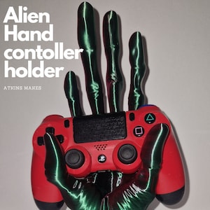 Könnte beinhalten: Eine 3D-gedruckte Alien-Hand hält einen roten PlayStation 4-Controller. Die Hand ist grün und hat lange, schlanke Finger. Die Basis der Hand ist schwarz und hat eine strukturierte Oberfläche. Der Text "Alien Hand controller holder" ist im Bild sichtbar.