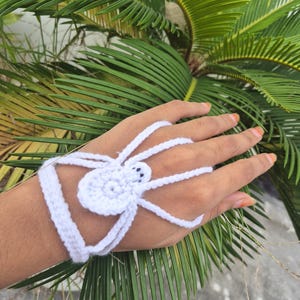 Puede incluir: Accesorio de mano de araña de ganchillo blanco. La araña tiene un cuerpo circular con ocho patas largas y dos pequeños ojos negros. El accesorio se usa en la mano y la muñeca, con correas que conectan la araña a la muñeca y los dedos.
