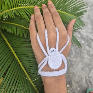 May include: A white crocheted hand accessory featuring a central spider motif with a round body and eight extended legs that loop around the fingers and wrist, creating a web-like structure that covers the back of the hand.