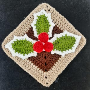 May include: A crocheted holly motif in a square shape, featuring green leaves with white edging, a brown center, and three red berries. The square is framed in a light beige colour, ideal for festive crafts.