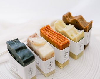 May include: Five bars of handmade soap in various colors, including black, white, orange, and brown, are arranged on a white plate. Each bar is wrapped in white paper with the text "Heart Soap" printed on it.