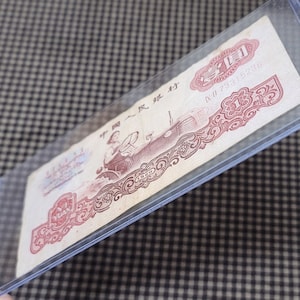 Può includere: Una banconota vintage della Repubblica Popolare Cinese, con un design rosso e nero con un ritratto di un uomo e il testo "中国人民银行" (Banca Popolare Cinese) e "贰拾圆" (Venti yuan).