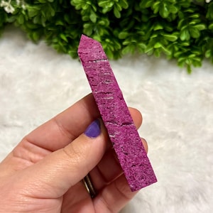 May include: A vibrant magenta-colored crystal point, rectangular in shape, with a textured surface. The crystal is held in a hand with a purple painted fingernail. The background features green foliage and a white, fluffy surface.