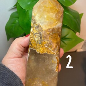 Puede incluir: Una punta de cristal de cuarzo transparente con inclusiones doradas. El cristal mide aproximadamente 15 cm de alto y tiene una forma natural y facetada.
