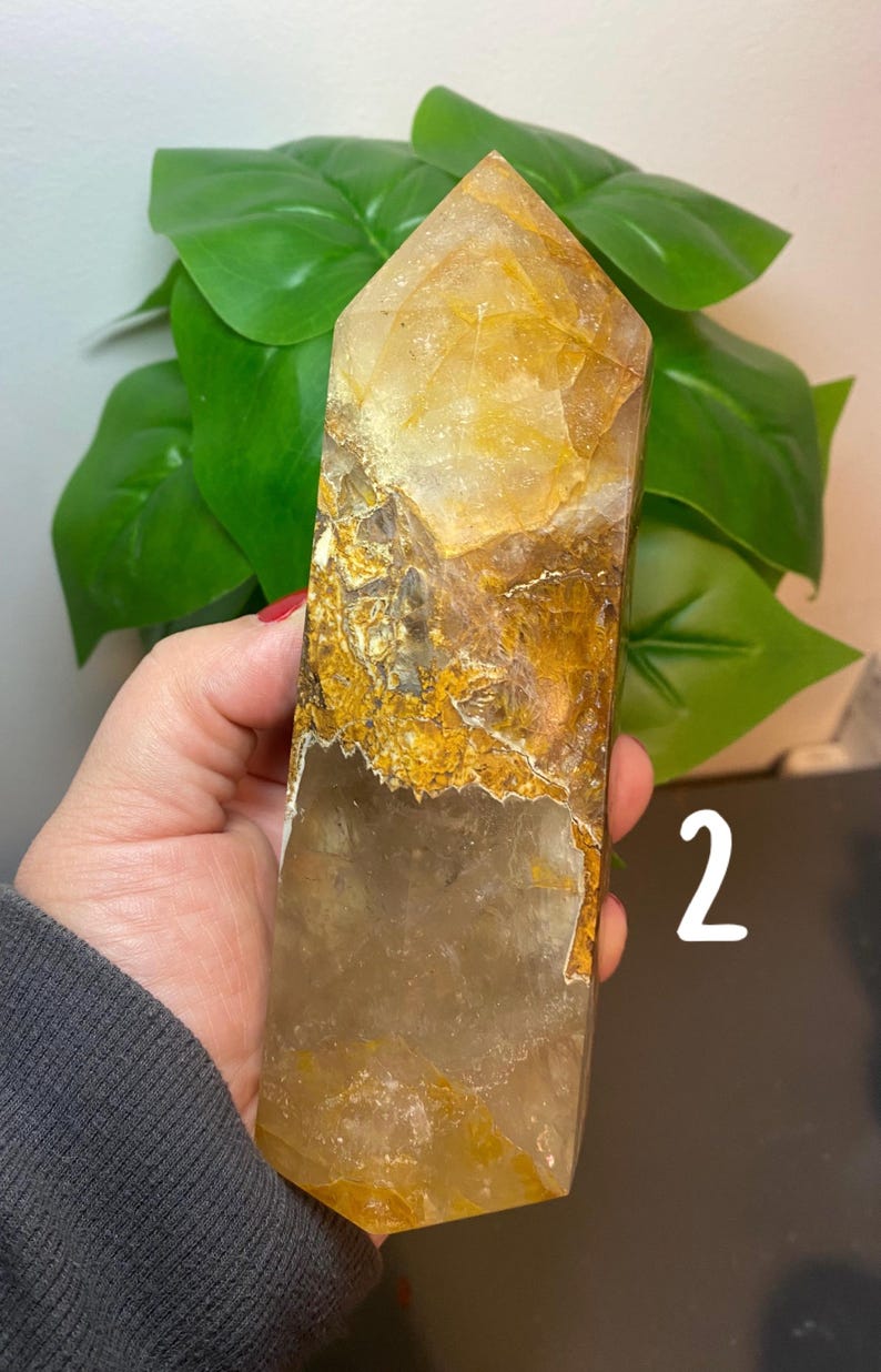 Puede incluir: Una punta de cristal de cuarzo transparente con inclusiones doradas. El cristal mide aproximadamente 15 cm de alto y tiene una forma natural y facetada.
