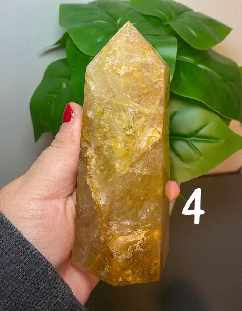 Puede incluir: Una gran punta de cristal de citrino amarillo claro con una superficie rugosa y texturizada. El cristal se sostiene en una mano, y el n&uacute;mero "4" est&aacute; escrito en el fondo.