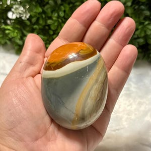May include: A polished, oval-shaped stone with a mix of colors, including orange, gray, tan, and white. The stone is smooth and appears to be held in a hand. The colors are layered in bands across the surface.