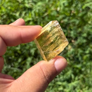 May include: A translucent, golden-yellow calcite crystal, held between fingers against a blurred green background. The crystal's facets catch the light, showcasing its natural texture and color. The crystal is a cube shape.