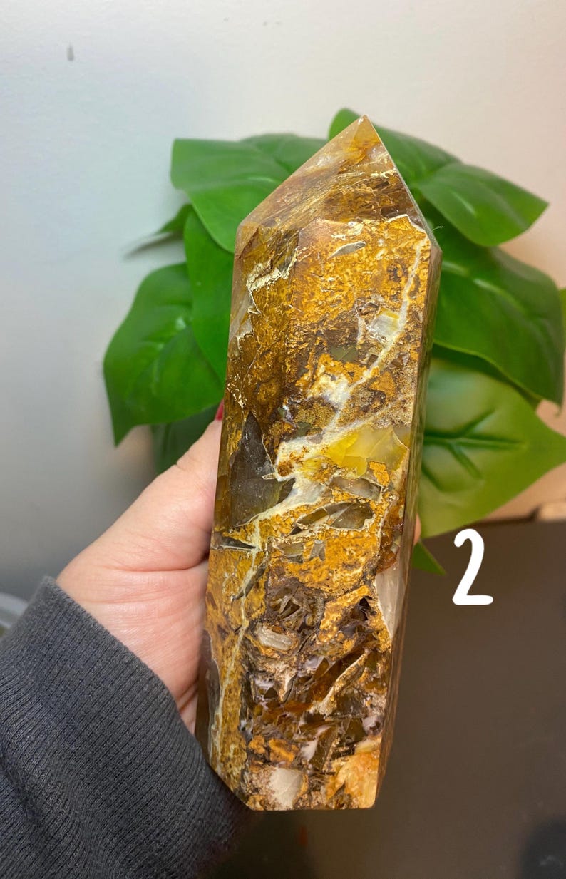 Puede incluir: Una gran punta de cristal de cuarzo de color marr&oacute;n dorado claro con inclusiones blancas y amarillas. El cristal mide aproximadamente 15 cm de alto y tiene una punta puntiaguda. El cristal est&aacute; sentado sobre una superficie marr&oacute;n.