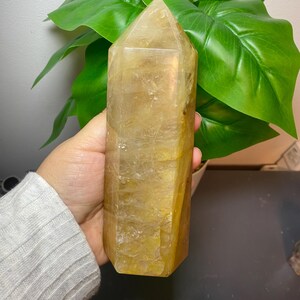 Puede incluir: Una gran punta de cristal de color amarillo dorado con facetas iridiscentes. El cristal es puntiagudo en la parte superior y tiene una base plana. Se est&aacute; sosteniendo en una mano.