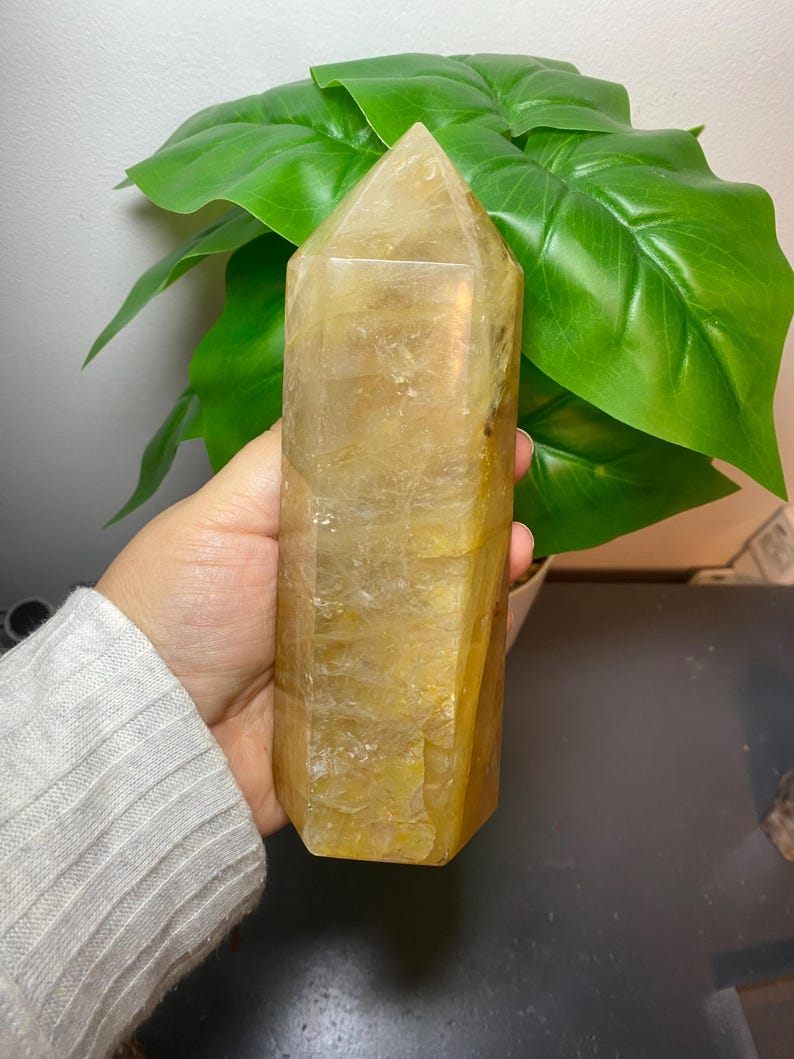 Puede incluir: Una gran punta de cristal de color amarillo dorado con facetas iridiscentes. El cristal es puntiagudo en la parte superior y tiene una base plana. Se est&aacute; sosteniendo en una mano.