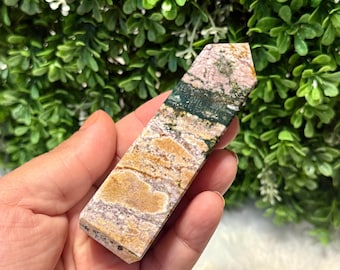 Puede incluir: Un obelisco rectangular de piedra preciosa pulida. La piedra presenta una mezcla de colores, incluyendo beige, rosa y verde, con un patrón natural y texturizado. El obelisco se sostiene en una mano sobre un fondo de follaje verde.