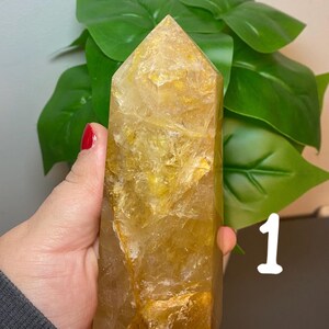 Puede incluir: Una gran punta de cristal de cuarzo transparente con un tono amarillo dorado. El cristal tiene una textura natural y rugosa y mide aproximadamente 15 cm de alto.