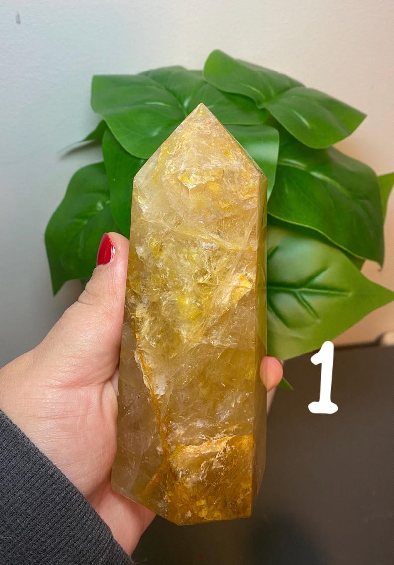 Puede incluir: Una gran punta de cristal de cuarzo transparente con un tono amarillo dorado. El cristal tiene una textura natural y rugosa y mide aproximadamente 15 cm de alto.