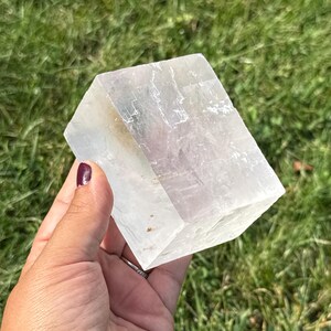 May include: A large, clear, rectangular crystal with a slightly translucent appearance. The crystal has a geometric shape and appears to be held against a backdrop of green grass. The crystal's edges are well-defined, and it has a natural, unpolished look.