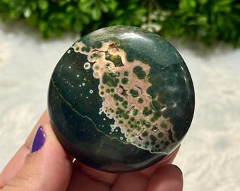 May include: A polished, round gemstone with a dark green base colour and a pattern of pink and white spots. The stone is held in a hand with a purple painted fingernail. The gemstone is a natural stone.