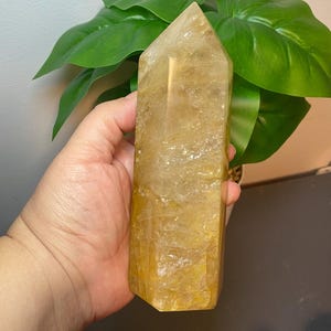 Puede incluir: Una gran punta de cristal de citrino amarillo claro con forma facetada. El cristal mide aproximadamente 15 cm de alto y tiene un aspecto natural y en bruto.