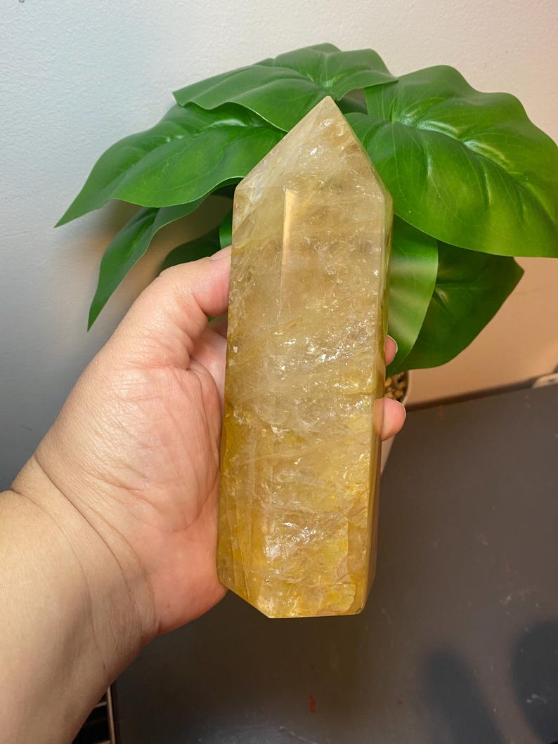 Puede incluir: Una gran punta de cristal de citrino amarillo claro con forma facetada. El cristal mide aproximadamente 15 cm de alto y tiene un aspecto natural y en bruto.