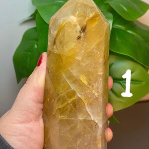 Puede incluir: Una gran punta de cristal de cuarzo transparente con inclusiones doradas. El cristal est&aacute; facetado y tiene una punta puntiaguda. El cristal est&aacute; siendo sostenido en una mano.