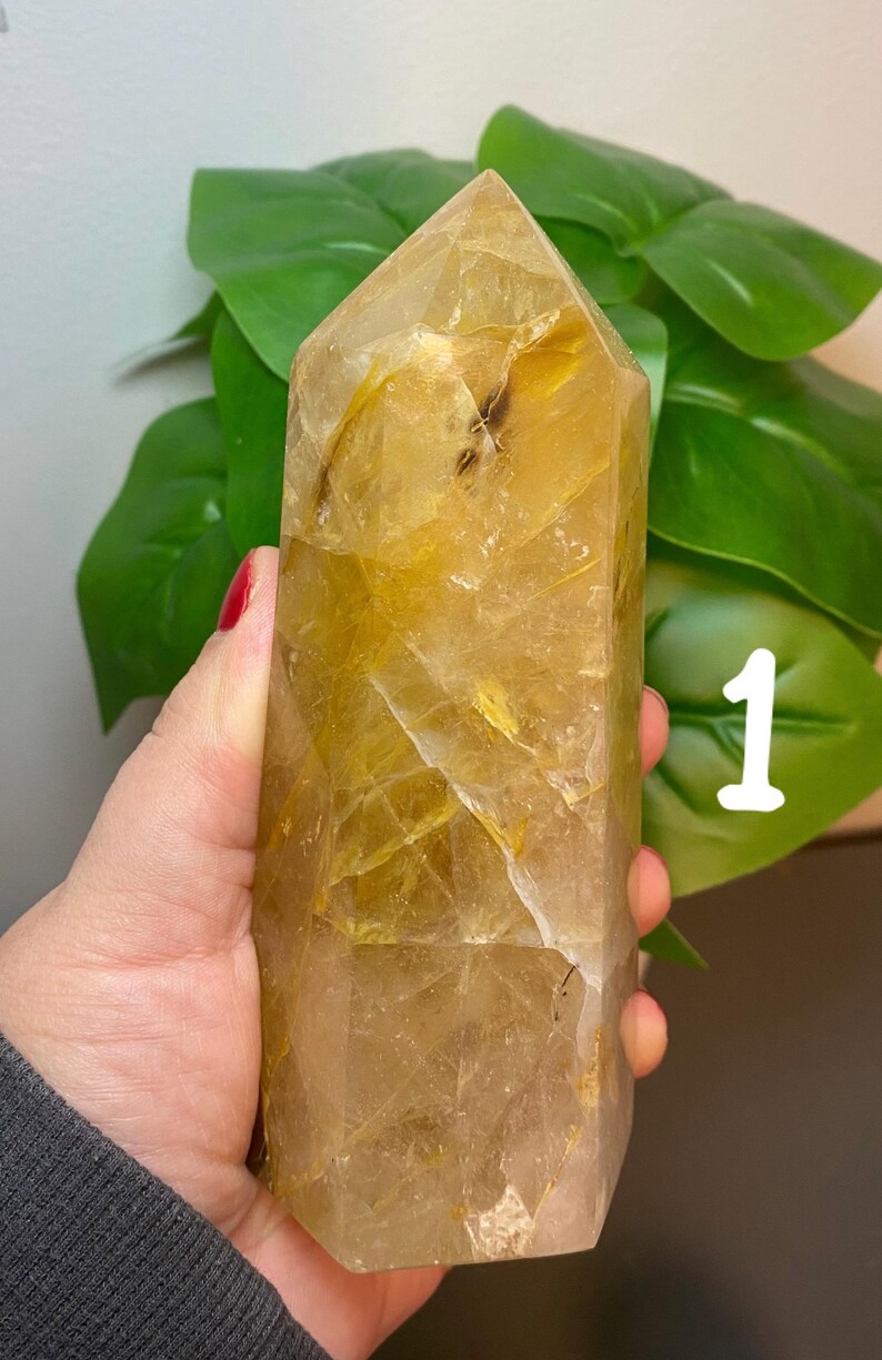 Puede incluir: Una gran punta de cristal de cuarzo transparente con inclusiones doradas. El cristal est&aacute; facetado y tiene una punta puntiaguda. El cristal est&aacute; siendo sostenido en una mano.