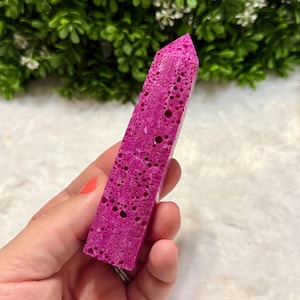 May include: A vibrant magenta crystal point with a porous texture. The crystal is held in a hand, showcasing its rectangular shape and pointed top. The background features green foliage.