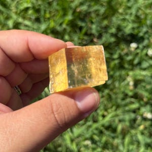 May include: A golden-yellow calcite crystal held between fingers against a blurred green background. The crystal is a rectangular shape with visible facets, catching the light and displaying a translucent quality. The crystal's color ranges from light to deep yellow.
