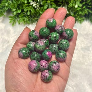 Puede incluir: Un conjunto de pequeñas piedras preciosas esféricas en tonos verdes y rosas. Las piedras pulidas se sostienen en una mano abierta, con un fondo verde borroso. Las piedras tienen un patrón moteado y probablemente se utilizan para joyería o decoración.